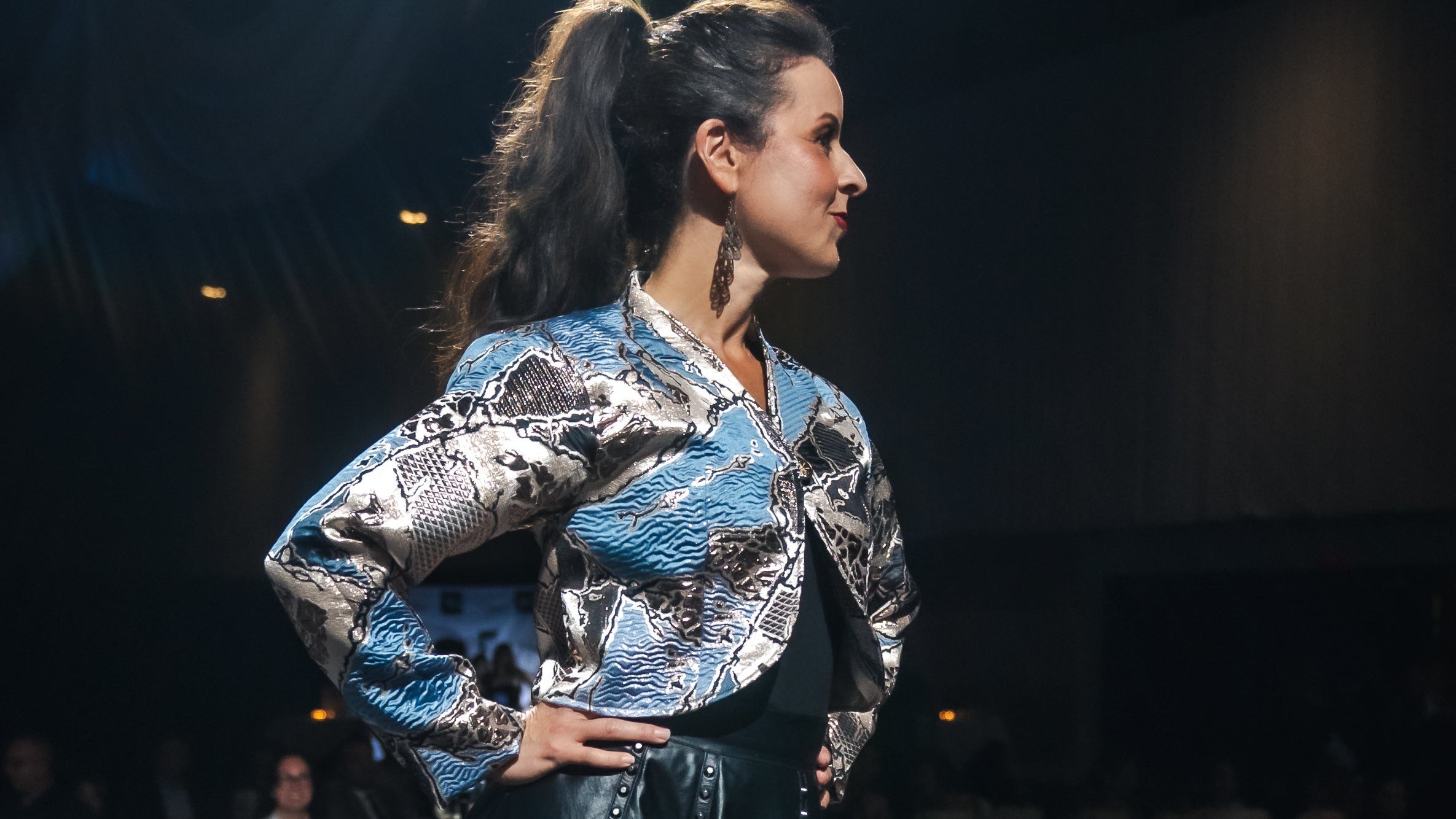 Model walking on a catwalk wearing a silver and blue jacket with a black leather skirt.