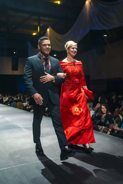 Man and woman walking on a fashion show runway, man in dark suit and woman in red dress.