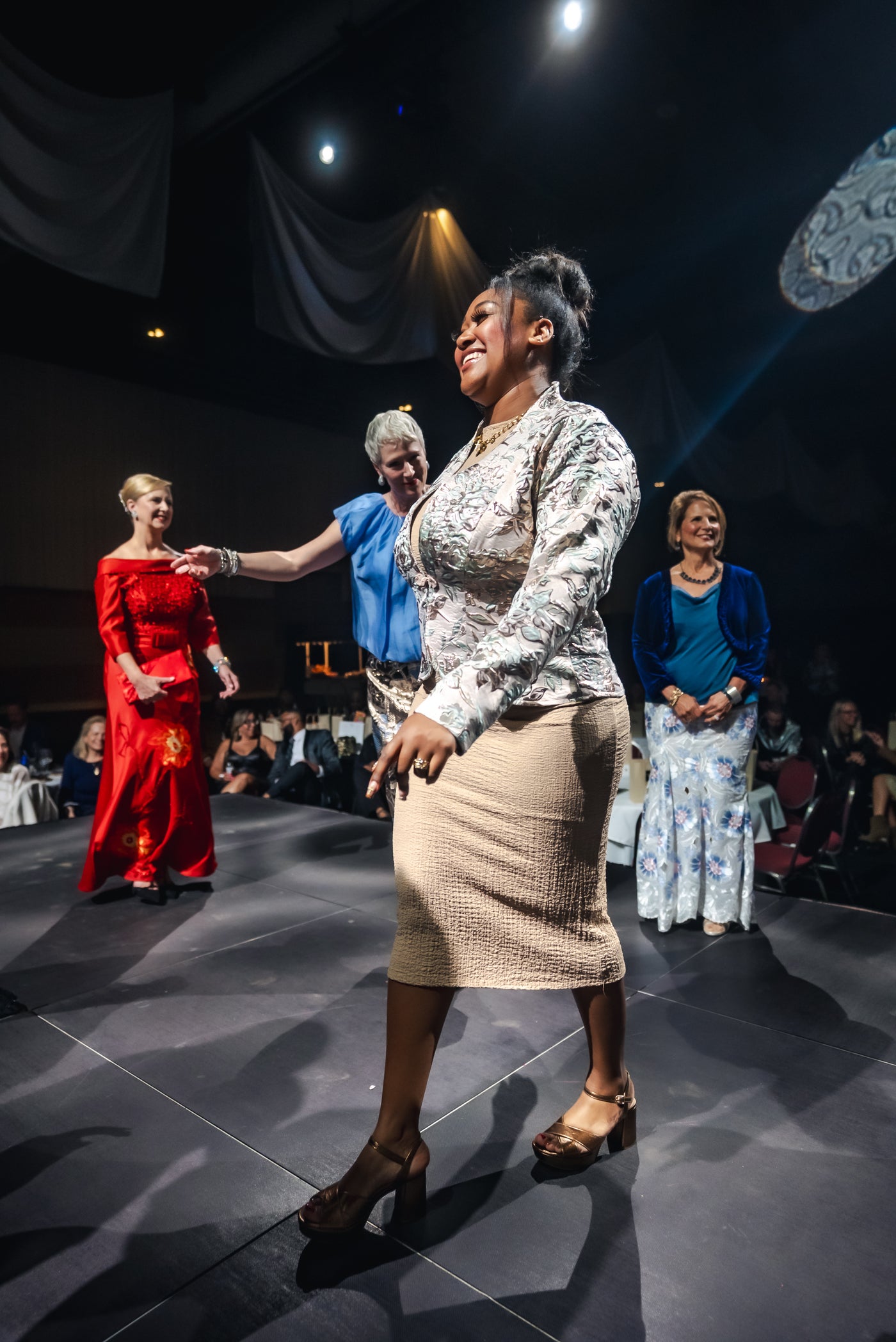 Woman in a patterned top and beige skirt on a stage with other people in formal attire.