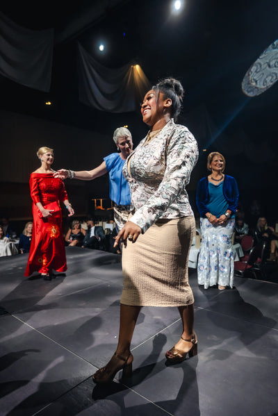 Woman in a patterned top and beige skirt on a stage with other people in formal attire.