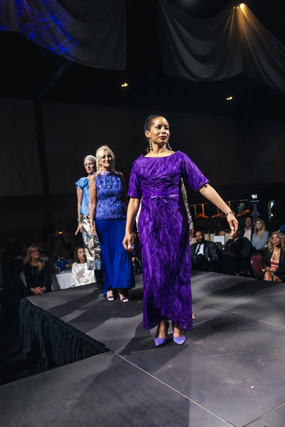 Model in a purple dress walking on a fashion show runway with an audience watching.