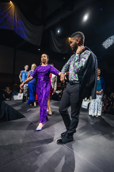 Model in a purple outfit walking on a runway with a man in a patterned shirt and dark pants beside her.